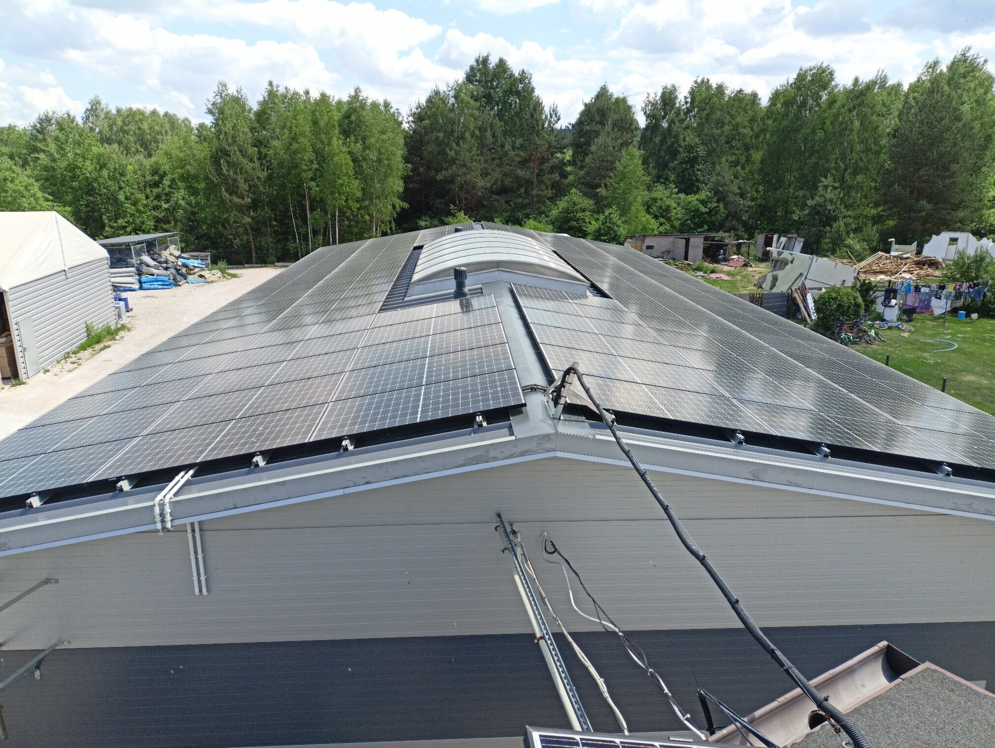 Widok z góry na dużą instalację paneli fotowoltaicznych na dachu budynku przemysłowego z lukarną, otoczonego zielenią, z widocznym sprzętem do czyszczenia paneli.