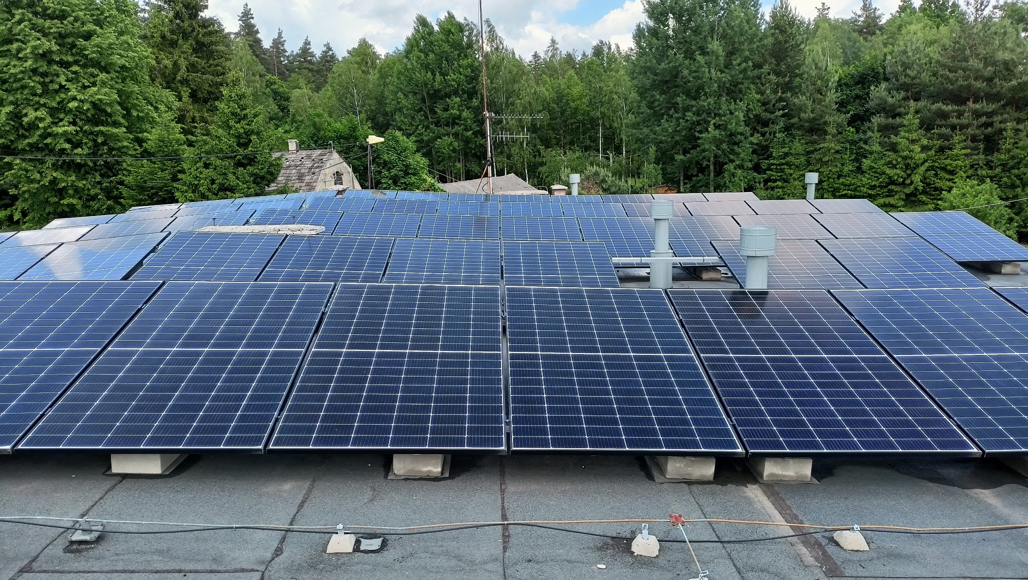 Widok z góry na panele fotowoltaiczne zamontowane na dachu budynku, z widocznymi kominami wentylacyjnymi i otaczającym lasem w tle.