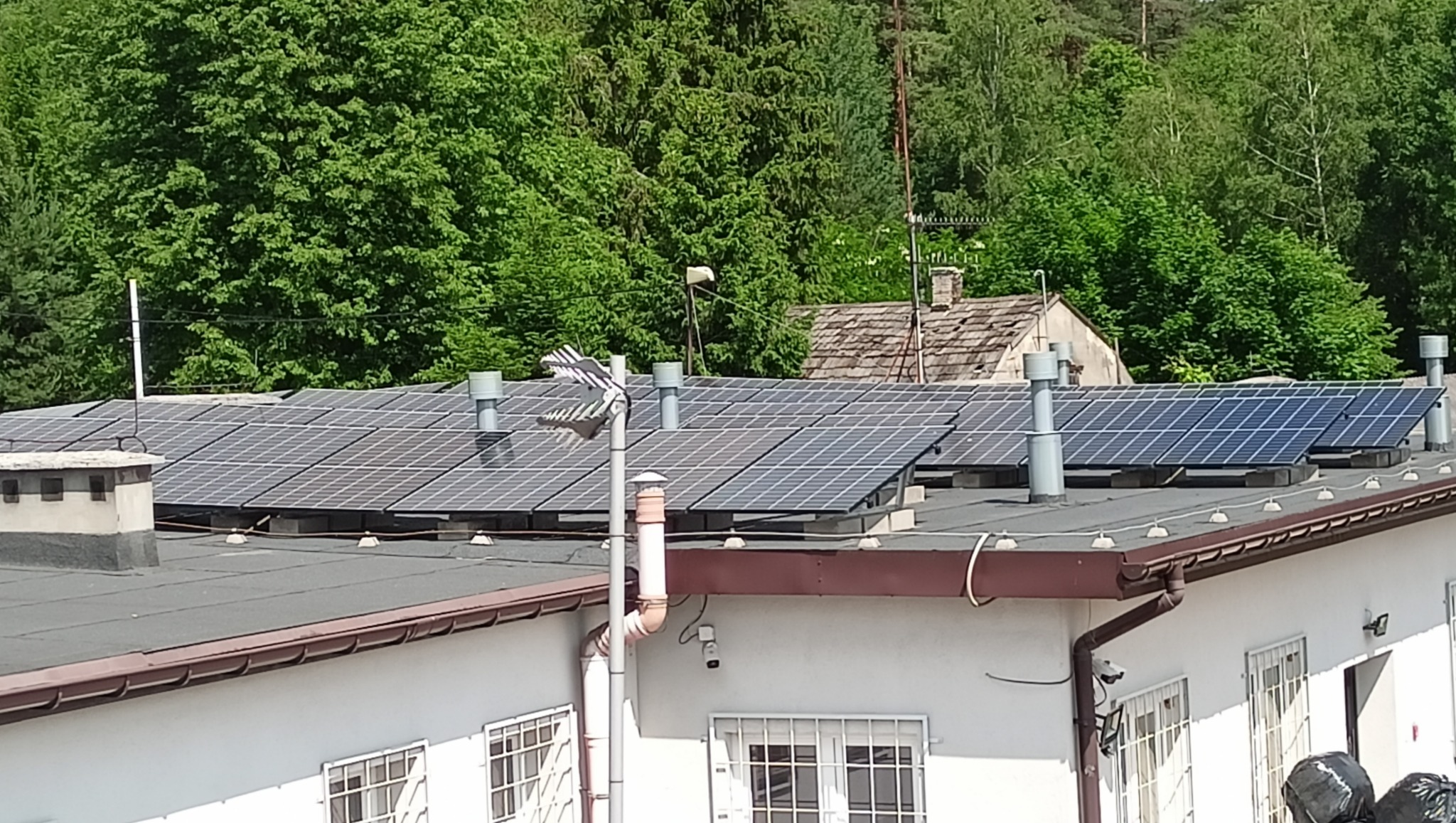 Panele fotowoltaiczne na dachu budynku z białymi ścianami i brązowym orynnowaniem, widoczne kominy wentylacyjne i drzewa w tle.