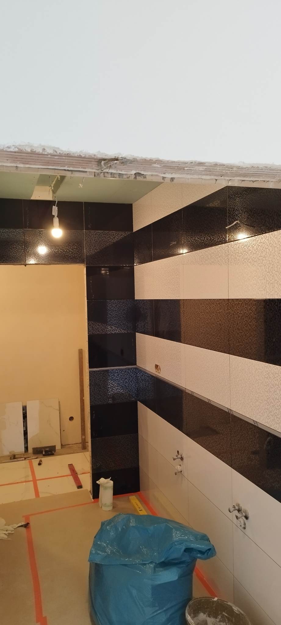 Częściowo wykończona łazienka z czarno-białymi płytkami ułożonymi w poziome pasy, widoczne niedokończone fugi, przygotowane podłoże pod płytki oraz worki na śmieci i narzędzia budowlane.