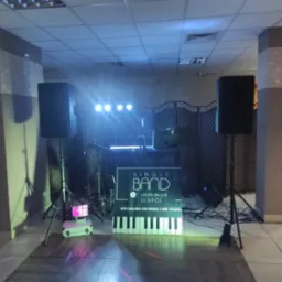 Scena z dwoma głośnikami na statywach, oświetleniem punktowym i banerem reklamowym zespołu muzycznego z klawiaturą fortepianu w tle kratownicy.