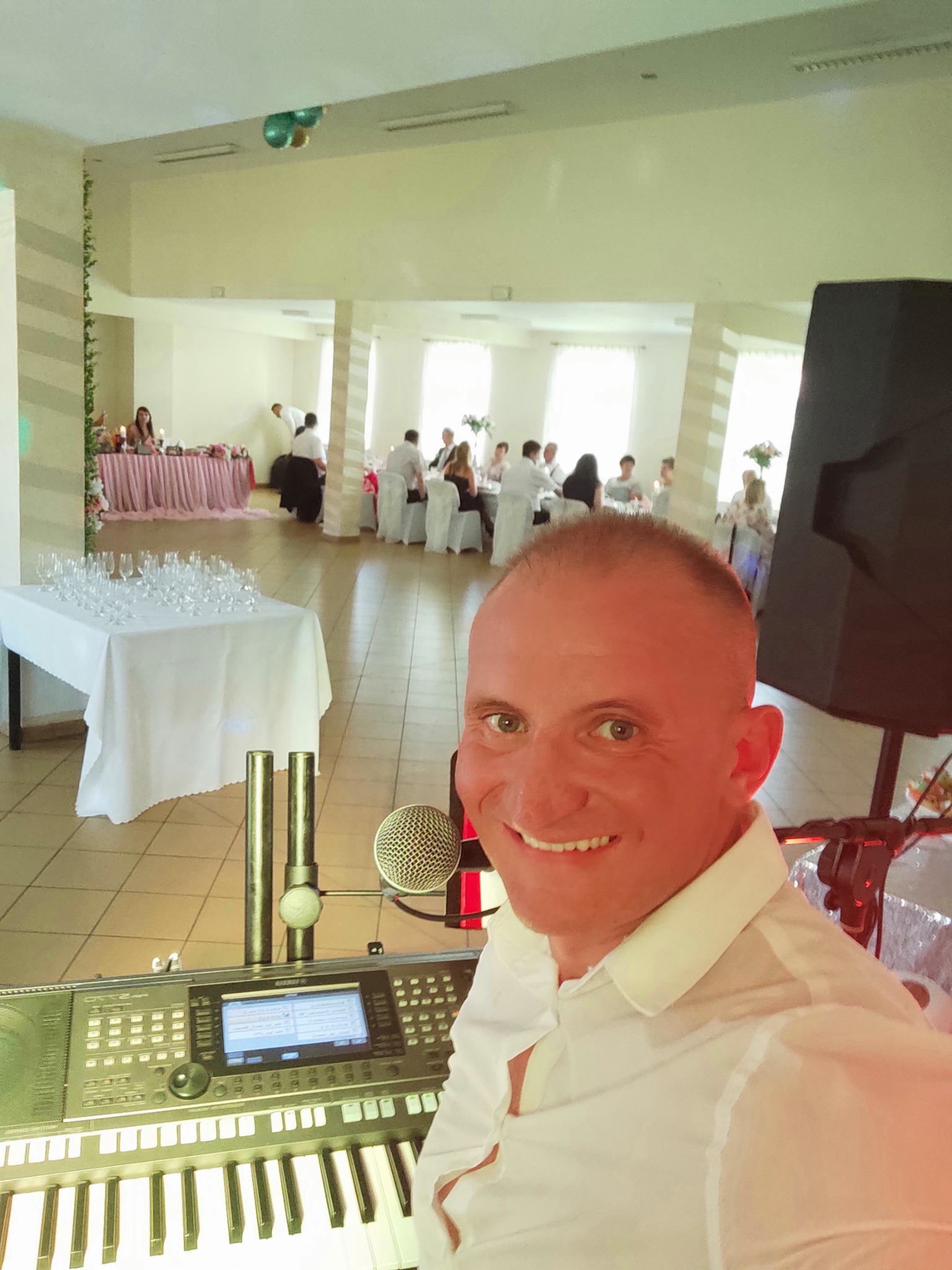 Muzyk z uśmiechem pozuje z keyboardem Yamaha Tyros na tle sali bankietowej z gośćmi weselnymi.