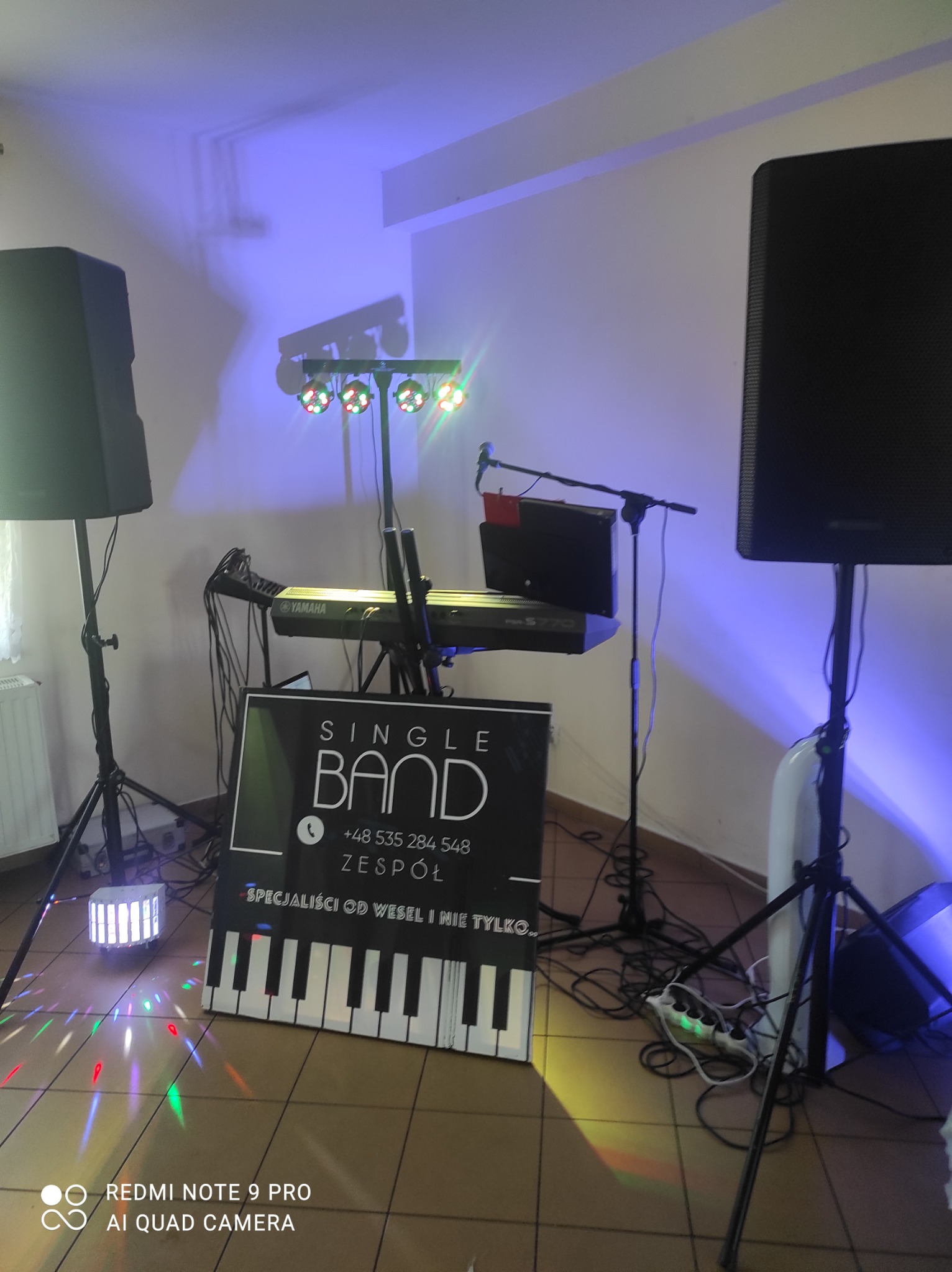 Sprzęt nagłośnieniowy i oświetleniowy na parkiecie, z widocznym keyboardem Yamaha, mikrofonami, kolumnami głośnikowymi i banerem reklamowym zespołu z klawiaturą fortepianu w tle, oświetlony...