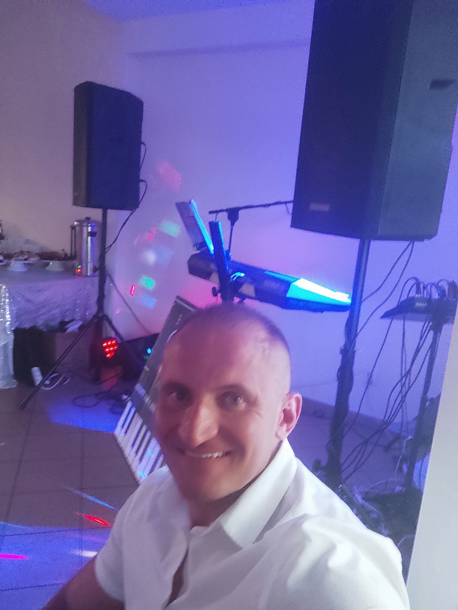 Uśmiechnięty mężczyzna robi selfie na tle sprzętu muzycznego: głośniki na statywach, klawisze i efekty świetlne w niebieskiej tonacji. W tle stolik z cateringiem.