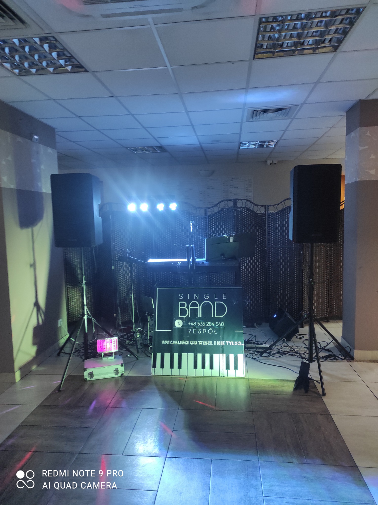 Scena z dwoma głośnikami na statywach, oświetleniem punktowym i banerem reklamowym zespołu muzycznego z klawiaturą fortepianu w tle kratownicy.