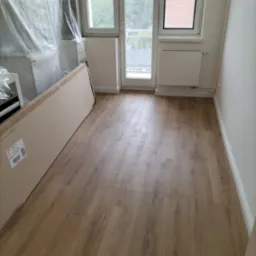 Ułożone panele podłogowe w jasnym odcieniu drewna, widoczne kartony z panelami i folia ochronna na meblach podczas prac wykończeniowych.