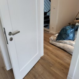 S.j bau - Otwarta biała rama drzwiowa z widoczną podłogą z paneli, workiem z gruzem i schodami w tle.