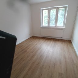 S.j bau - Puste pomieszczenie z panelami podłogowymi w odcieniach brązu, białymi ścianami i oknem z widokiem na zieleń. Świeże, wykończone wnętrze z grzejnikiem pod oknem i widocznymi gniazdkami elektrycznymi.