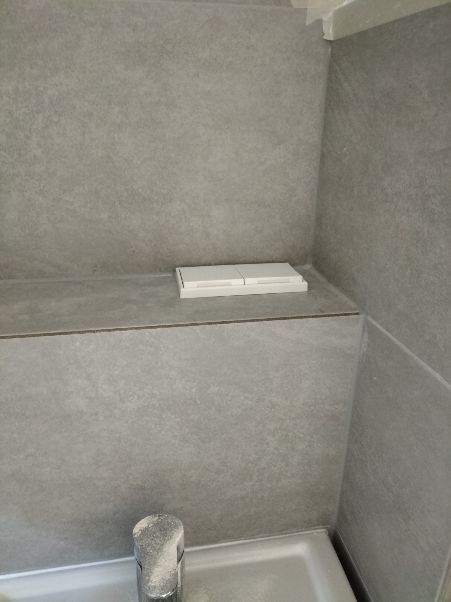 Szare płytki w łazience, widoczny fragment baterii umywalkowej z osadem oraz przycisk spłuczki zabudowany w ścianie.