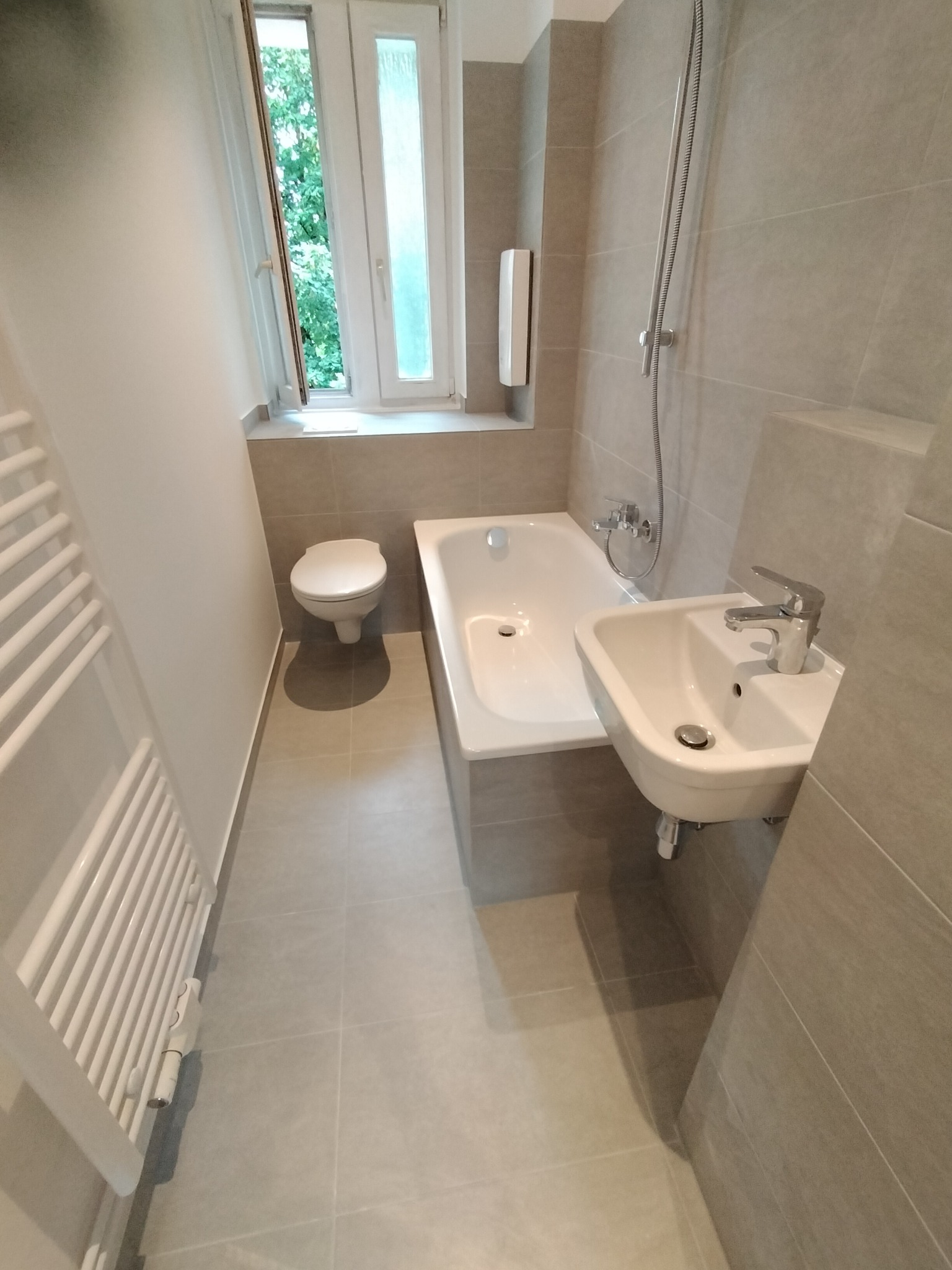 Jasna łazienka z szarymi płytkami, białym wyposażeniem i grzejnikiem drabinkowym, widok na okno z zielenią na zewnątrz.