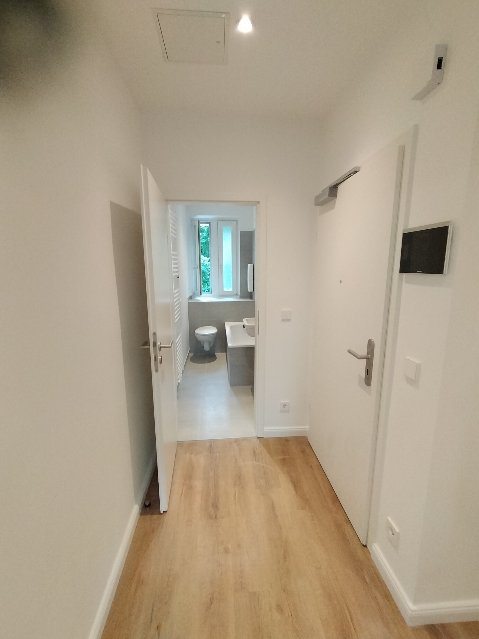 Korytarz z panelami podłogowymi prowadzący do nowocześnie wykończonej łazienki z widoczną umywalką i toaletą, oświetlony punktowym światłem.