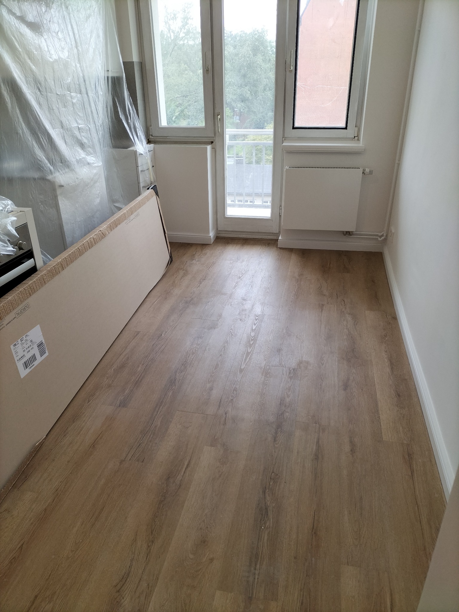 Ułożone panele podłogowe w jasnym odcieniu drewna, widoczne kartony z panelami i folia ochronna na meblach podczas prac wykończeniowych.