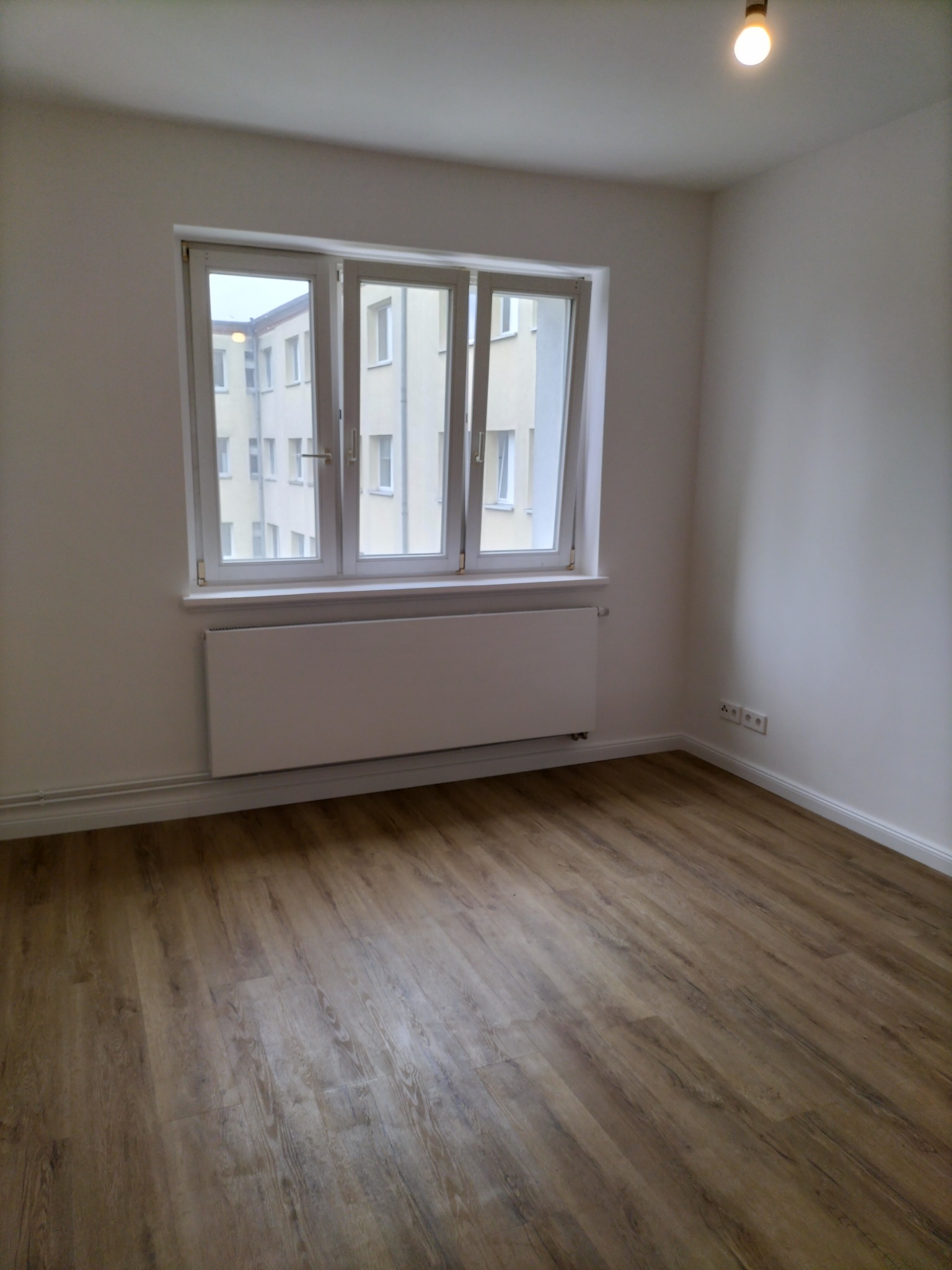 Pusty pokój z panelami podłogowymi w odcieniach brązu, białymi ścianami, grzejnikiem pod oknem i widokiem na budynek z zewnątrz.