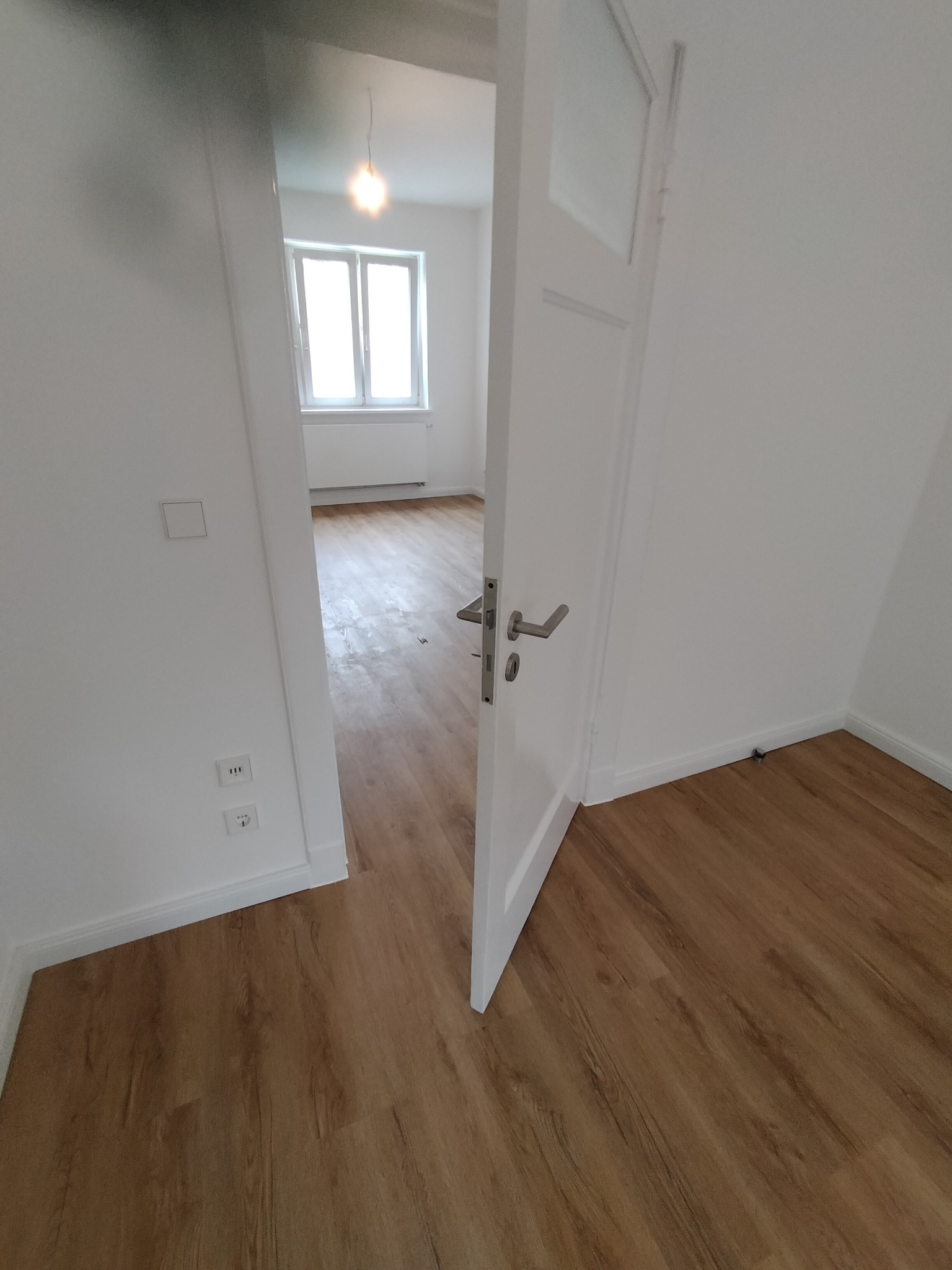 Jasne panele podłogowe w nowym mieszkaniu widoczne przez uchylone białe drzwi z szybą; w tle okno z grzejnikiem.