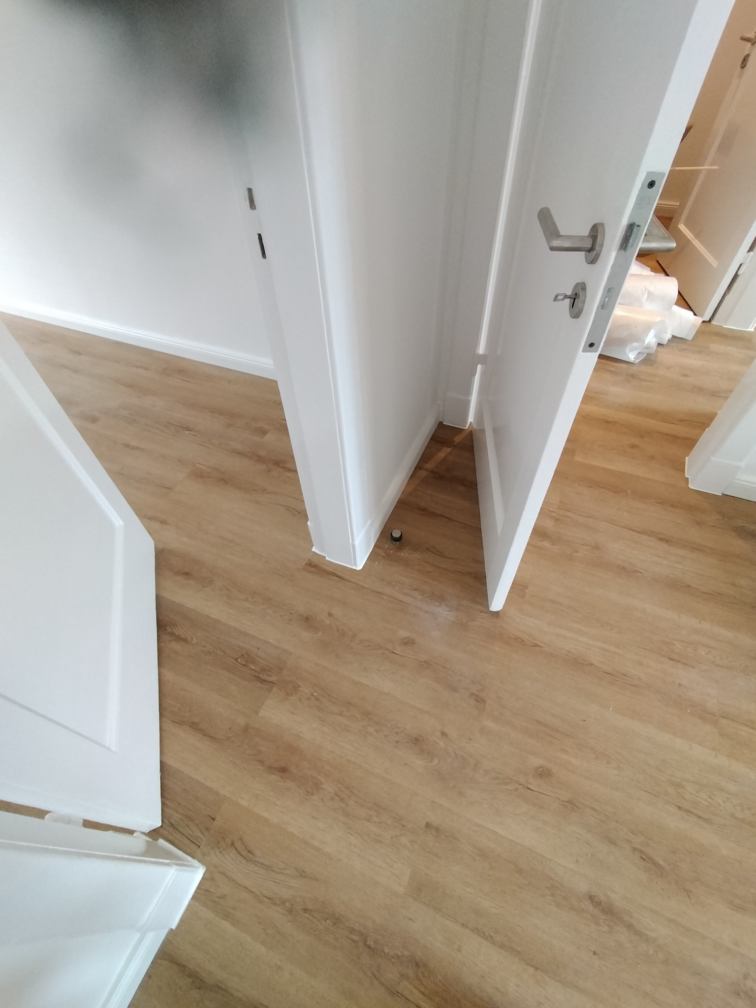 Jasne panele podłogowe imitujące drewno, białe drzwi wewnętrzne z srebrną klamką, widoczne białe listwy przypodłogowe i fragmenty folii ochronnej na podłodze w tle.