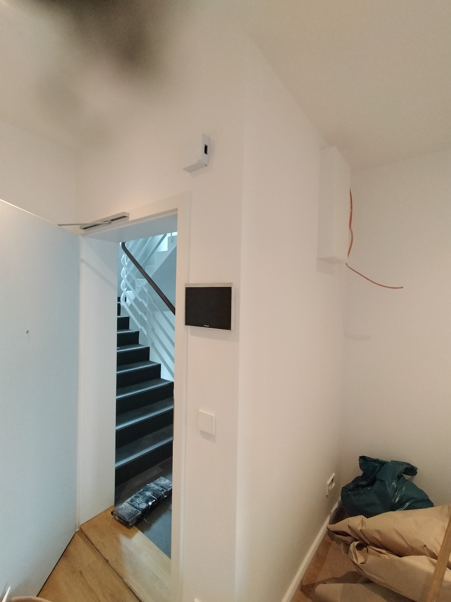 Białe wnętrze w trakcie wykończenia: widoczne schody, domofon, wystające kable i worki z materiałami budowlanymi w rogu.
