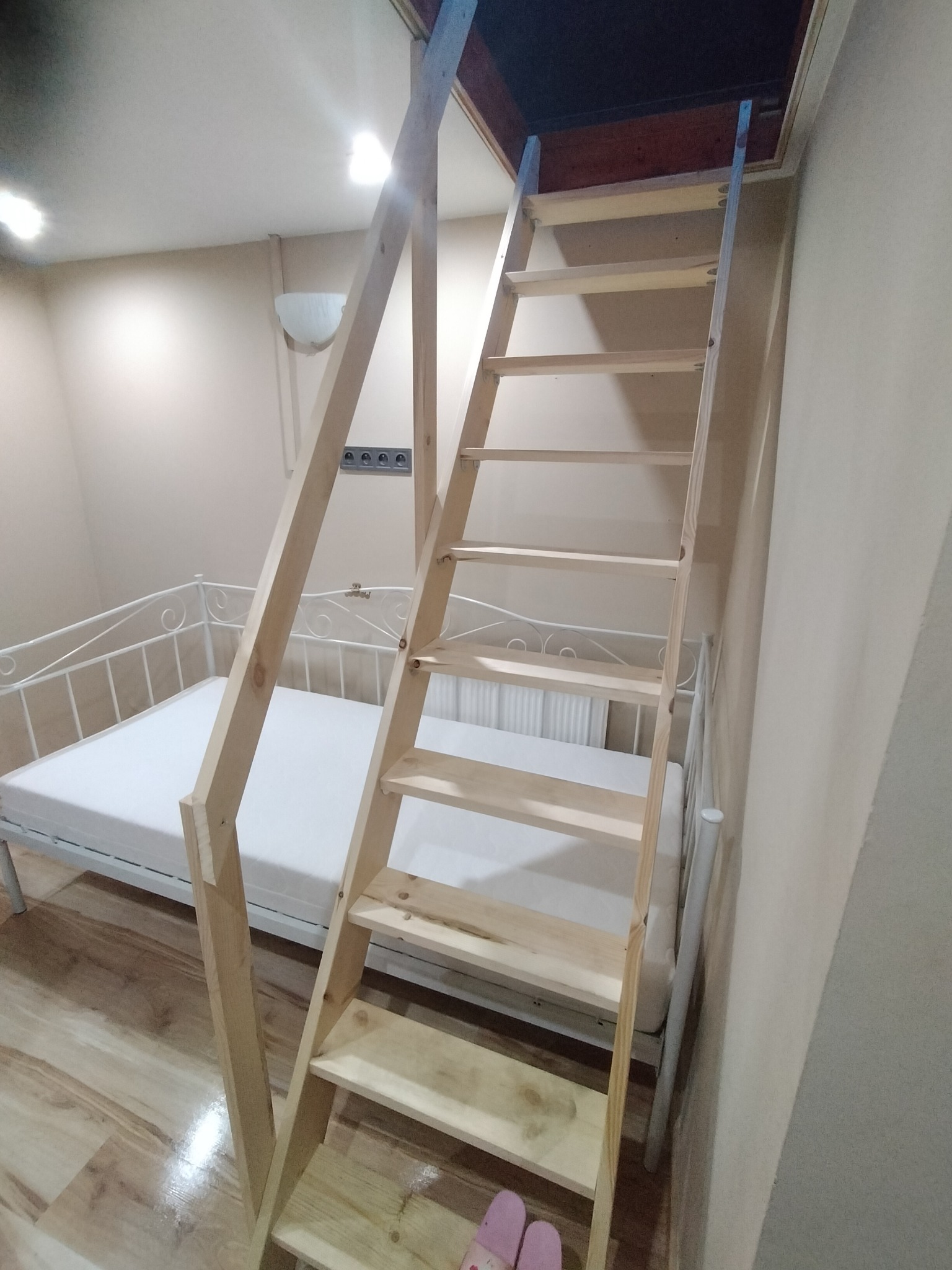 Drewniane schody strychowe prowadzące do otworu w suficie, umieszczone obok białego łóżka z metalową ramą w pokoju o jasnych ścianach.