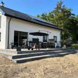 Nowoczesny dom z białymi ścianami i ciemną dachówką, panele słoneczne na dachu, taras z szarej kostki brukowej w trakcie budowy, teren wokół wyrównany, widoczny grill i meble ogrodowe.