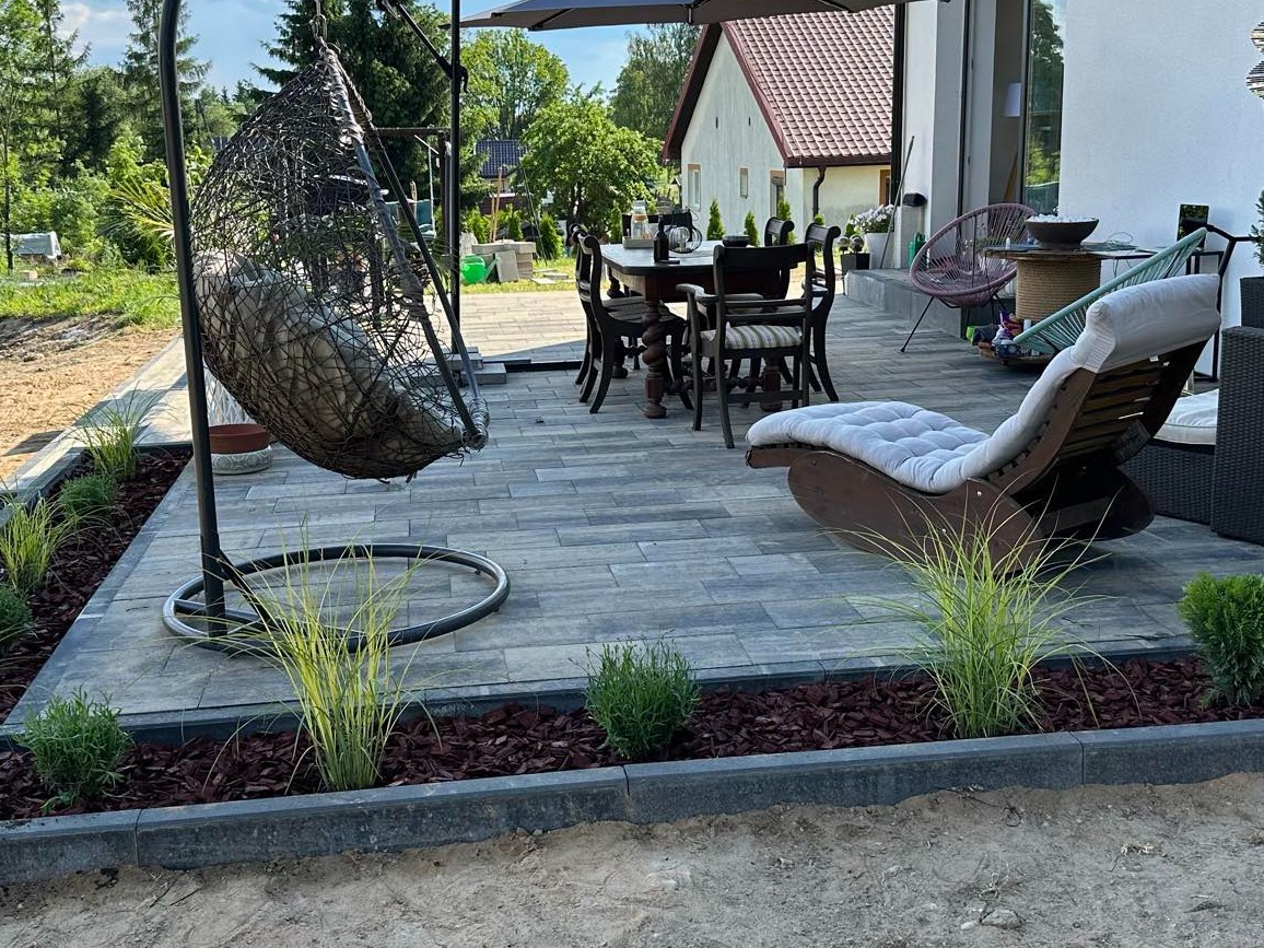 Szara kostka brukowa na tarasie z meblami ogrodowymi, hamakiem wiszącym i rabatami z roślinami ozdobnymi, widok z poziomu gruntu.