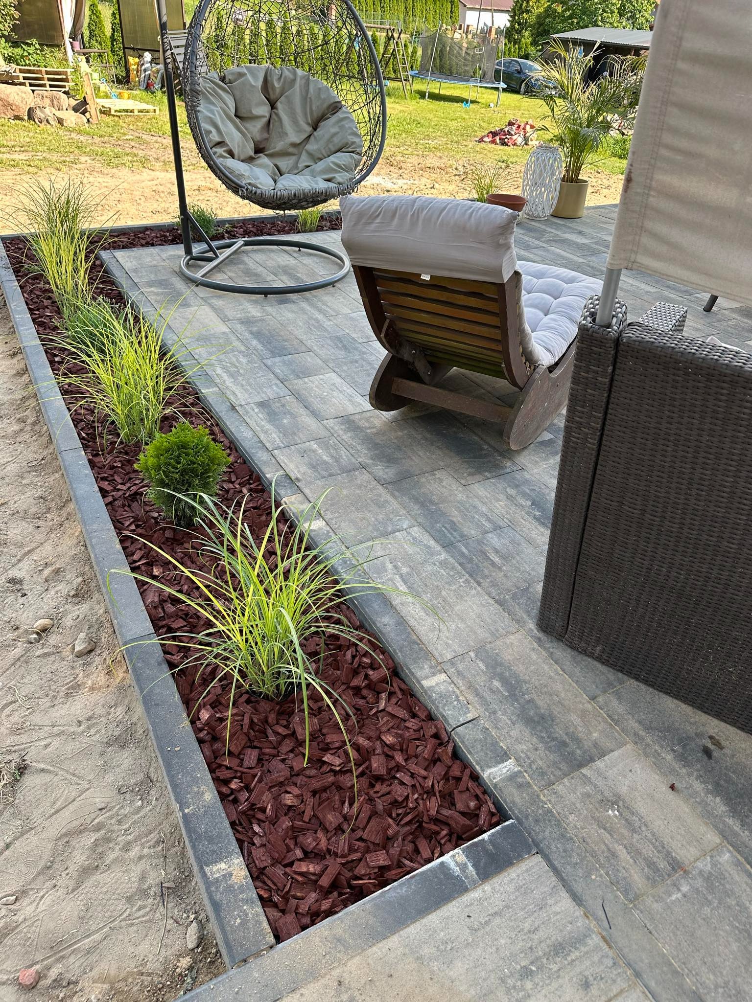 Szare płyty chodnikowe tworzące patio z rattanowymi meblami, wiszącym fotelem i rabatą z ozdobnymi trawami i czerwoną korą.