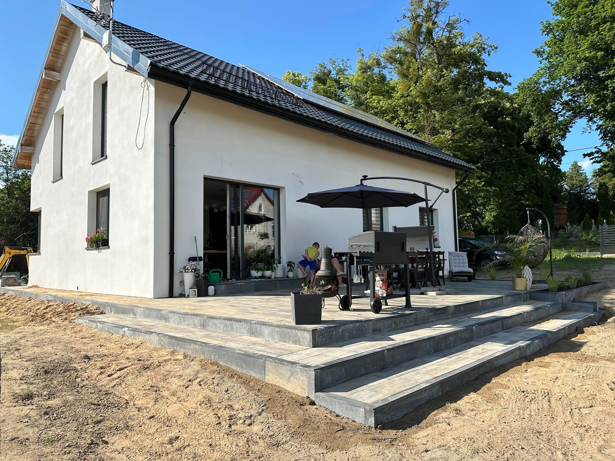 Nowoczesny dom z białymi ścianami i ciemną dachówką, taras wyłożony jasnoszarą kostką brukową z ciemnoszarymi obrzeżami, częściowo otoczony piaskiem, na tarasie grill, parasol, meble ogrodowe...