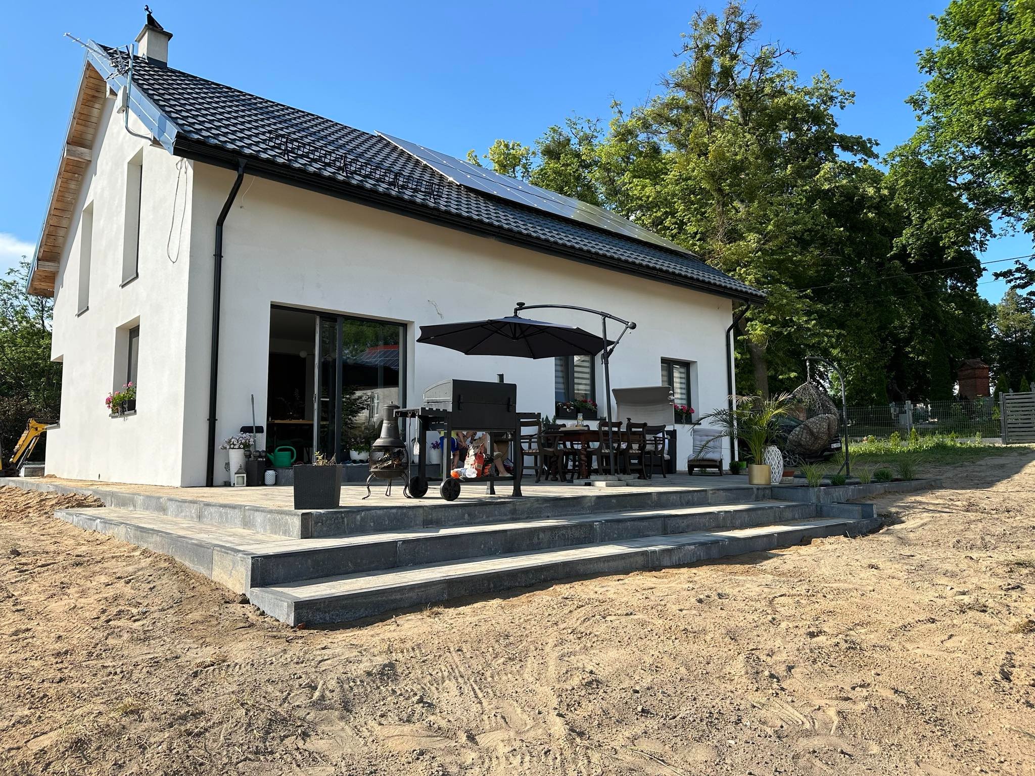 Nowoczesny dom z białymi ścianami i ciemną dachówką, panele słoneczne na dachu, taras z szarej kostki brukowej w trakcie budowy, teren wokół wyrównany, widoczny grill i meble ogrodowe.