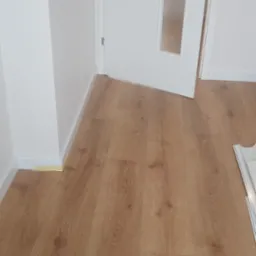 Jasnobrązowe panele podłogowe ułożone w pokoju z białą ścianą i częściowo otwartymi białymi drzwiami z szybą; widoczny fragment listwy przypodłogowej przy ścianie oraz resztki taśmy malarskiej.