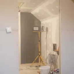 Poddasze w trakcie wykańczania: widoczne płyty kartonowo-gipsowe na ścianach i suficie, szpachlowane łączenia, reflektor na trójnogu oświetlający szarą ścianę, rolka wełny mineralnej na podłodze.