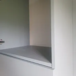 Wnętrze szafki z półką, widoczne zawiasy. Świeżo pomalowane, szare wnętrze szafki kontrastuje z białymi drzwiami. Detal wykończenia wnętrza.