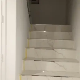 Wykończone schody z białego marmuru z metalowymi listwami na krawędziach stopni, zabezpieczone żółtą taśmą malarską, z szklaną balustradą i widocznym fragmentem instalacji elektrycznej na ścianie.