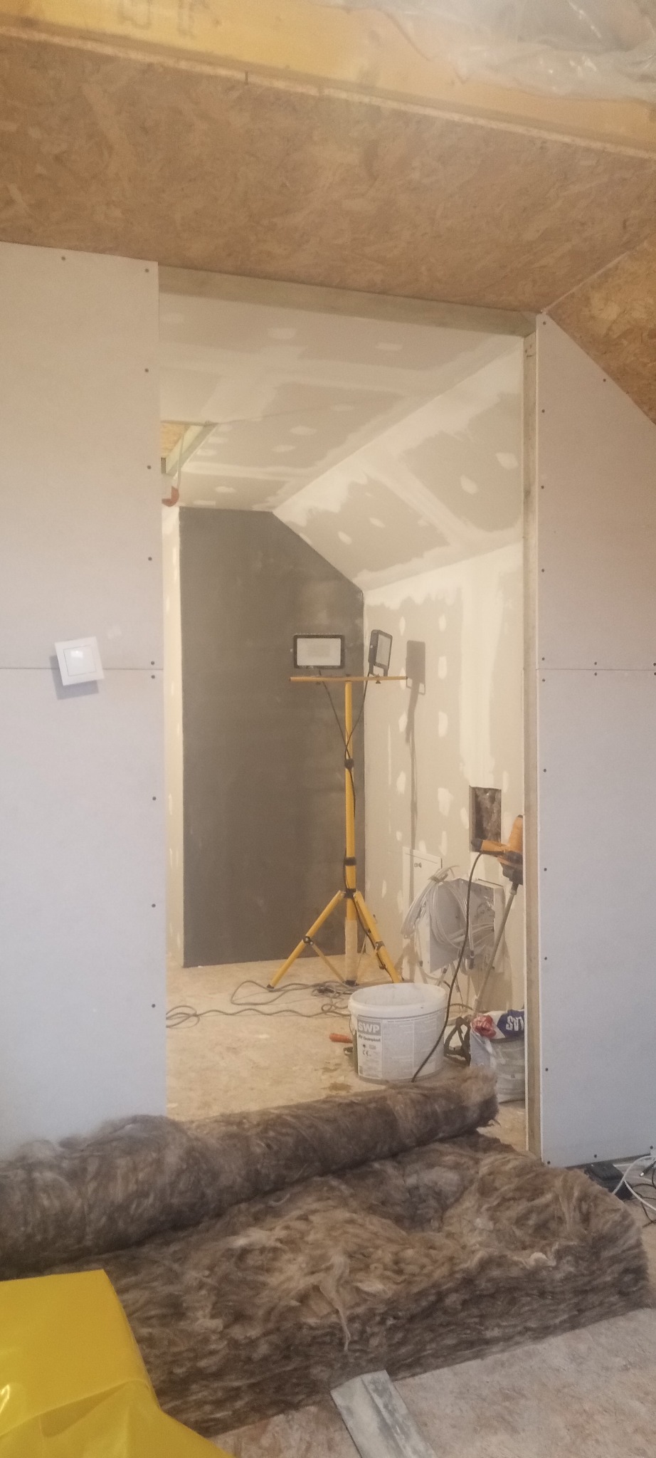 Poddasze w trakcie wykańczania: widoczne płyty kartonowo-gipsowe na ścianach i suficie, szpachlowane łączenia, reflektor na trójnogu oświetlający szarą ścianę, rolka wełny mineralnej na podłodze.