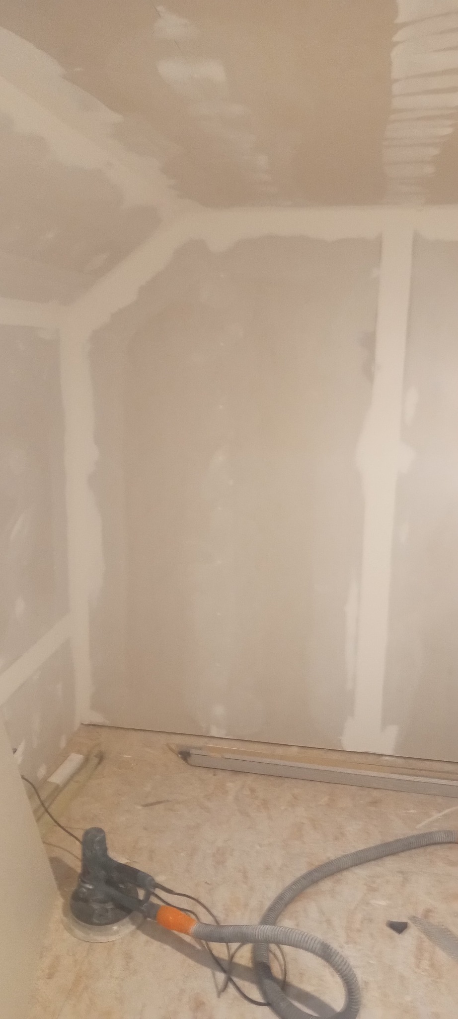 Wnętrze pokoju w trakcie remontu, ściany pokryte płytami kartonowo-gipsowymi, widoczne ślady szpachlowania, na podłodze szlifierka oscylacyjna z podłączonym odkurzaczem.