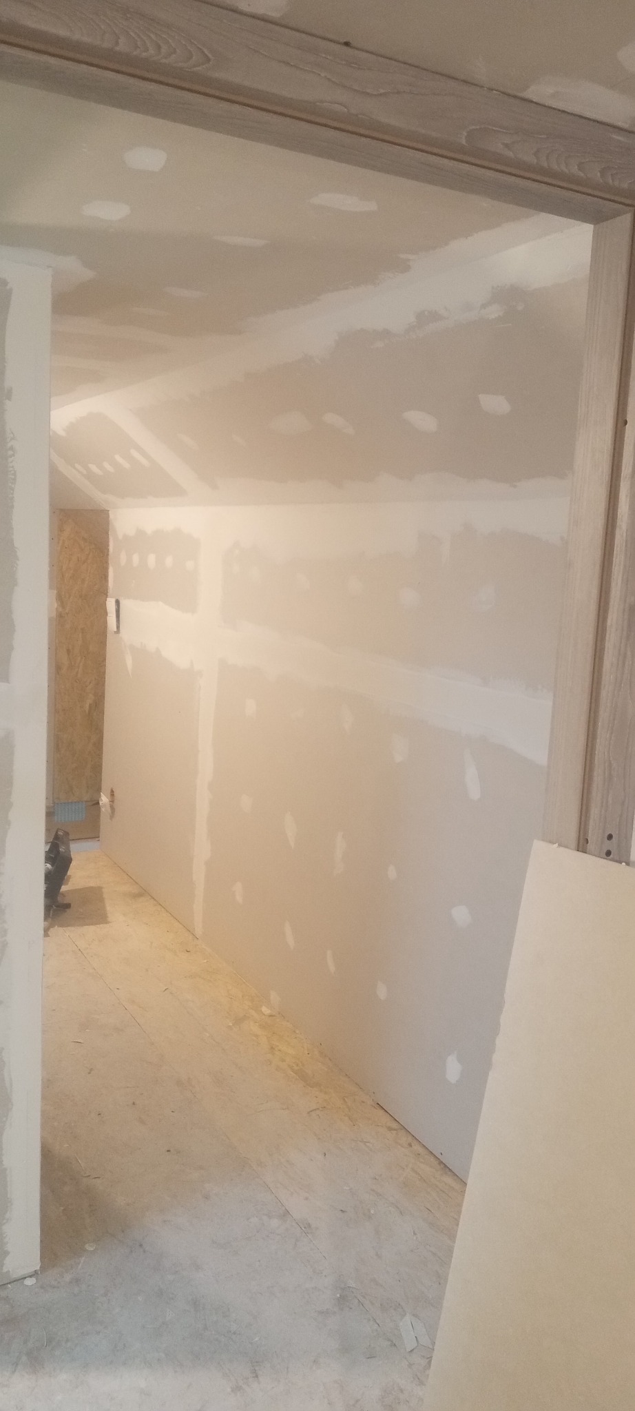 Wnętrze w trakcie prac wykończeniowych: ściany z płyt kartonowo-gipsowych ze śladami szpachlowania, widoczne łączenia płyt i miejsca po wkrętach wypełnione masą, surowa drewniana podłoga, fragment...