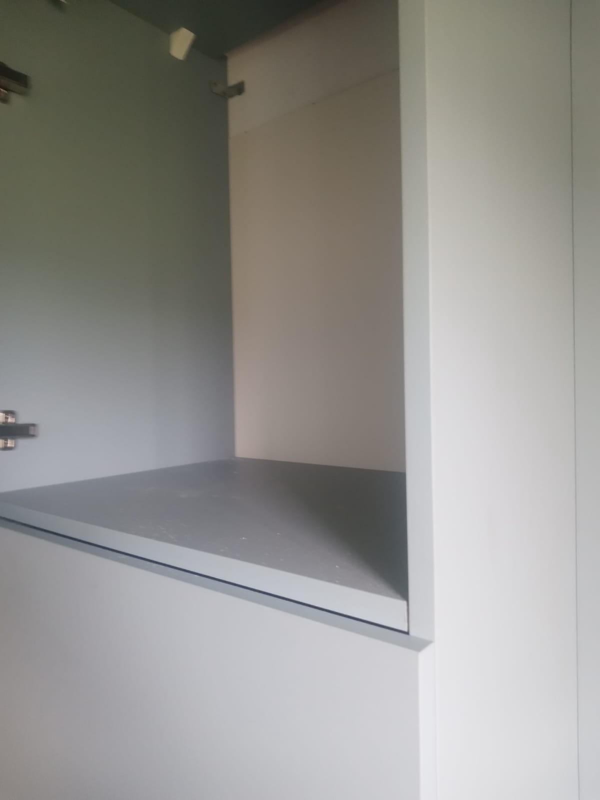 Wnętrze szafki z półką, widoczne zawiasy. Świeżo pomalowane, szare wnętrze szafki kontrastuje z białymi drzwiami. Detal wykończenia wnętrza.