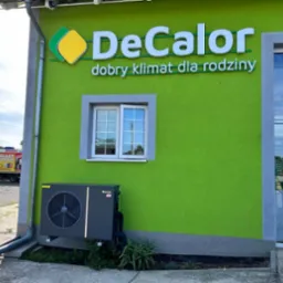Szara pompa ciepła zamontowana na zielonej ścianie budynku z oknem i logo firmy DeCalor, na zewnątrz widoczny samochód dostawczy z napisem Pellet Ekogroszek.