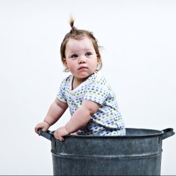 Karolina Bukowska - Niemowlę z zadumanym wyrazem twarzy siedzi w metalowej balii na tle białego studia.