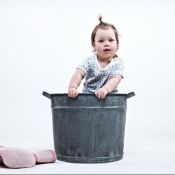 Karolina Bukowska - Uśmiechnięte dziecko z małym kucykiem w koszulce w gwiazdki, wystające z metalowego wiadra na tle białego studia, obok leży pluszowa zabawka.