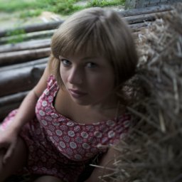 Karolina Bukowska - Portret kobiety w kwiecistej sukience, siedzącej na tle drewnianych bali i siana, z delikatnym, naturalnym oświetleniem i lekko rozmytym tłem.