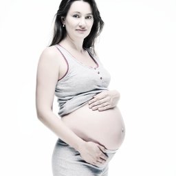 Karolina Bukowska - Portret kobiety w ciąży w szarym komplecie dresowym na białym tle, dłonie ułożone na brzuchu, subtelny uśmiech.