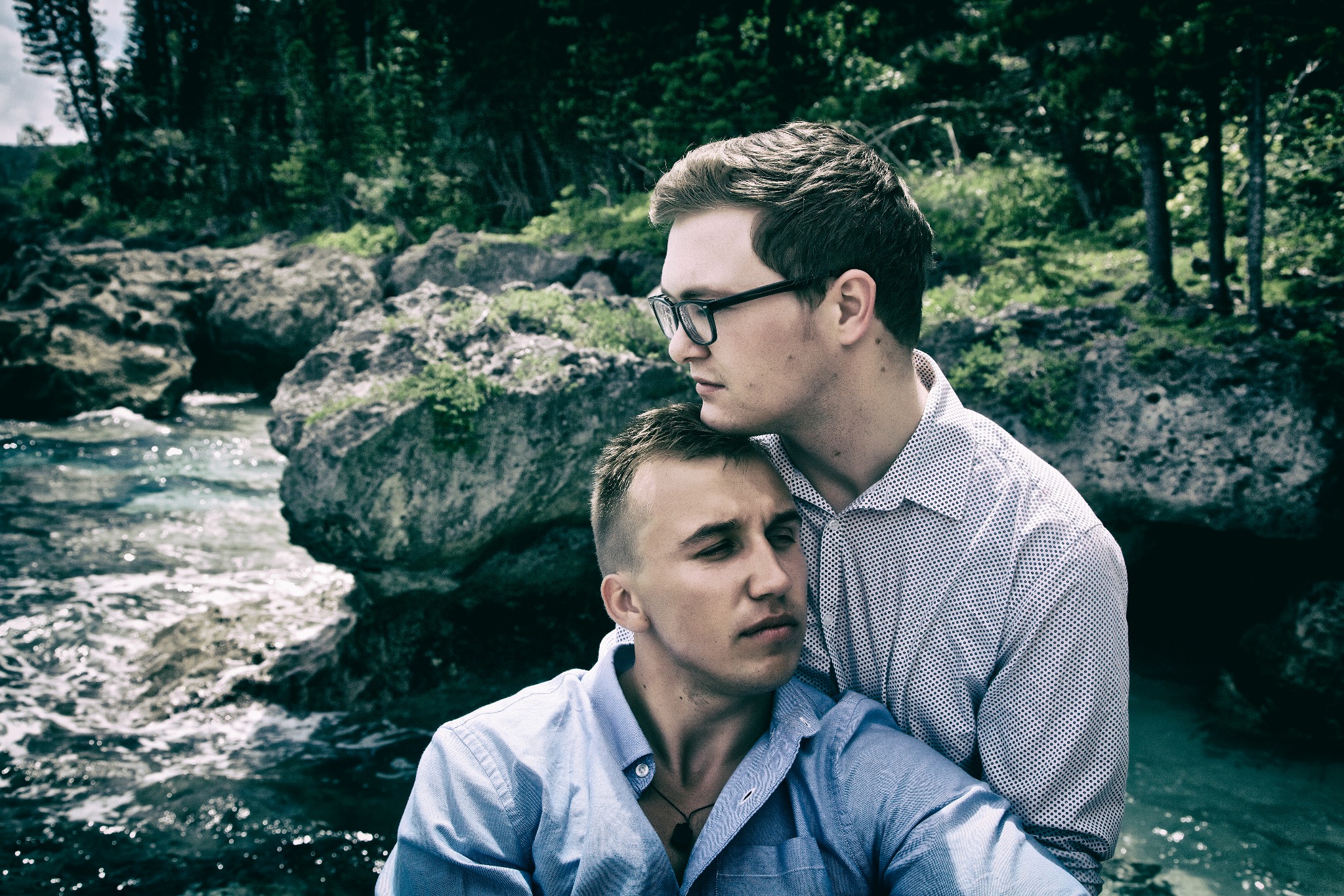 Dwóch mężczyzn w koszulach pozuje na tle skalistego krajobrazu z wodą i zielenią, jeden oparty głową o drugiego.