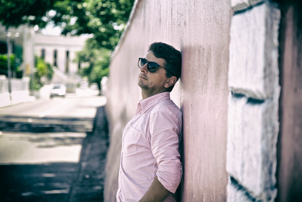 Mężczyzna w okularach przeciwsłonecznych, oparty o różową ścianę w plenerze miejskim, w tle budynek z kolumnami i zieleń drzew.