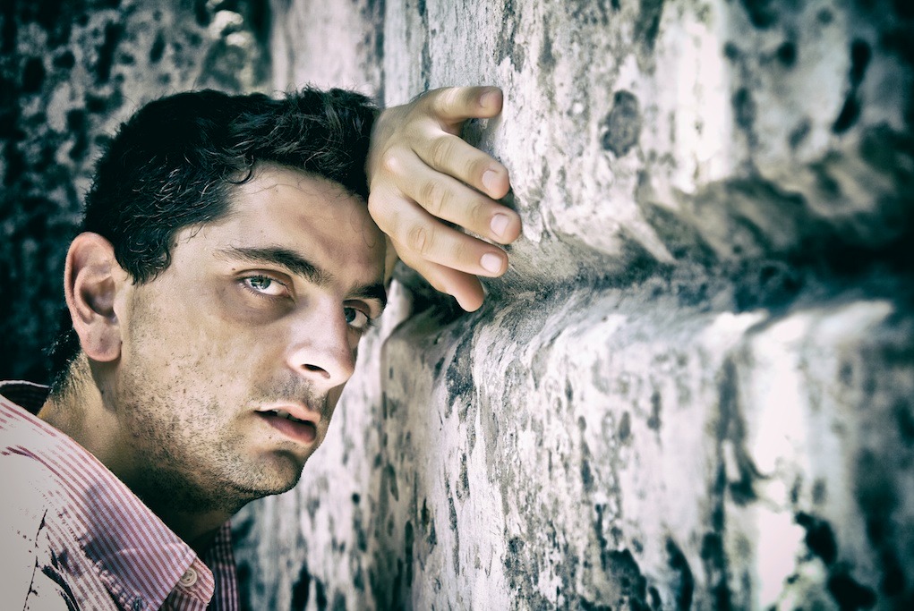 Portret młodego mężczyzny opierającego się o szorstką, kamienną ścianę w plenerze, z ręką dotykającą kamienia, w naturalnym świetle i zamyślonym spojrzeniem.