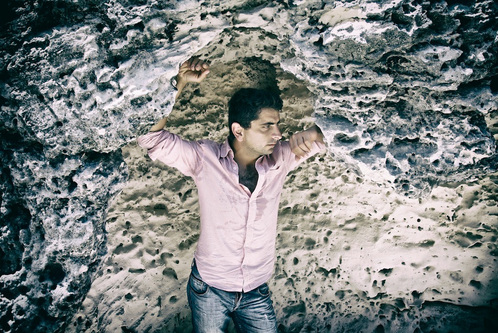 Mężczyzna w różowej koszuli pozuje oparty o porowatą, skalną ścianę w plenerze, z rękami uniesionymi ku górze, spoglądając w bok.