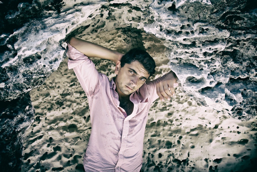 Mężczyzna w rozpiętej koszuli w paski pozuje w skalnej wnęce, z rękami opartymi o nierówne ściany.