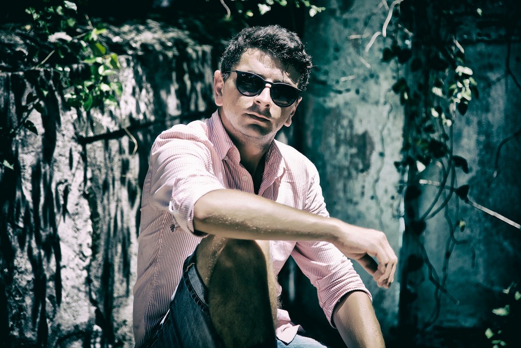 Mężczyzna w okularach przeciwsłonecznych i różowej koszuli w paski, siedzący w plenerze na tle kamiennej ściany porośniętej bluszczem.