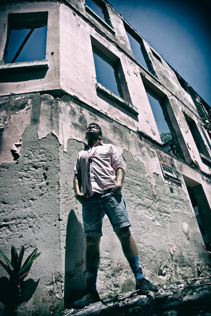 Mężczyzna w różowej koszuli i dżinsowych szortach stoi przed zrujnowanym budynkiem z pustymi oknami, spoglądając w górę, z rękami w kieszeniach, w plenerze.