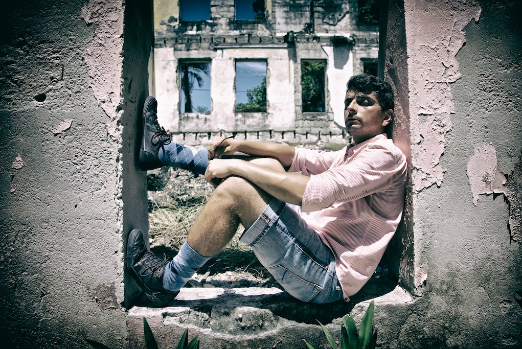 Mężczyzna w różowej koszuli siedzi w oknie zniszczonego budynku, nosi dżinsowe szorty i skarpetki w romby, a w tle widoczne okna z odbiciami drzew.