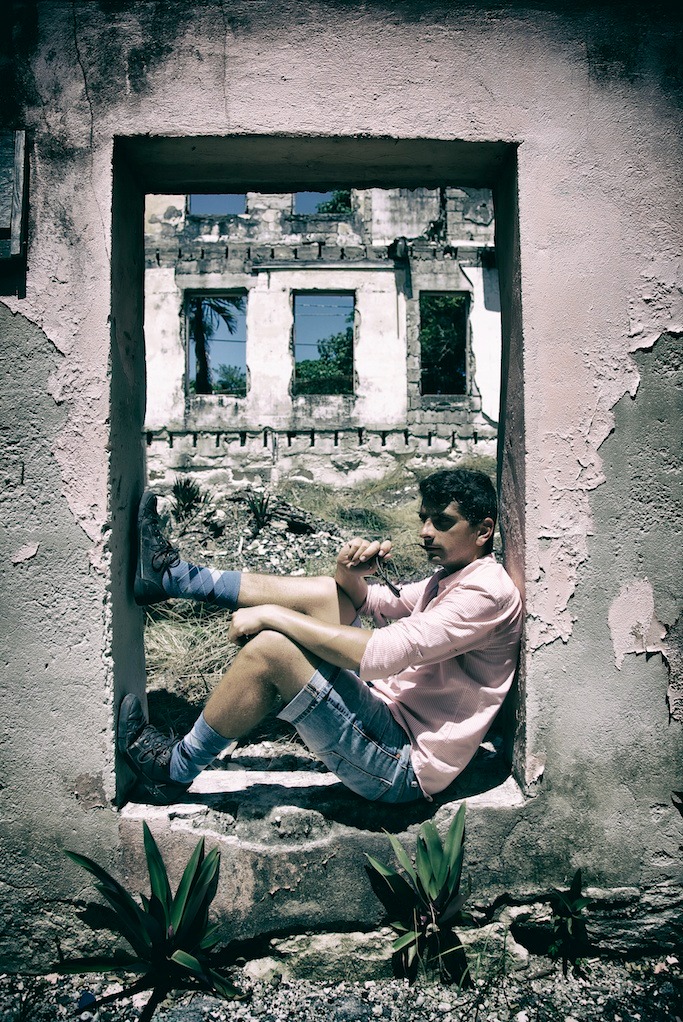 Mężczyzna w różowej koszuli i dżinsowych spodenkach siedzi w zniszczonym oknie budynku, trzymając okulary w ręku, z widokiem na budynek z oknami odbijającymi niebo i roślinność.