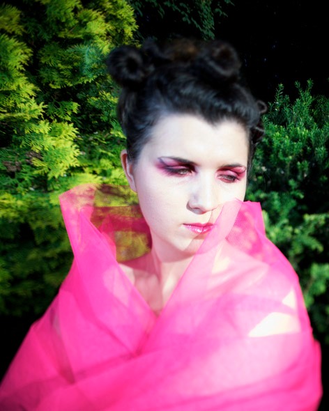 Portret kobiety w plenerze, z intensywnym różowym makijażem oczu i ust, owiniętej w jaskrawą, różową, przezroczystą tkaninę, na tle zielonych krzewów.