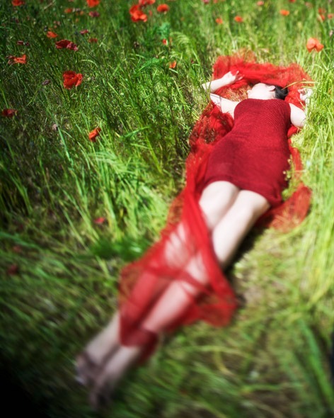 Kobieta w czerwonej sukience leży na łące z makami, otulona czerwoną tkaniną, z rękami uniesionymi nad głową.